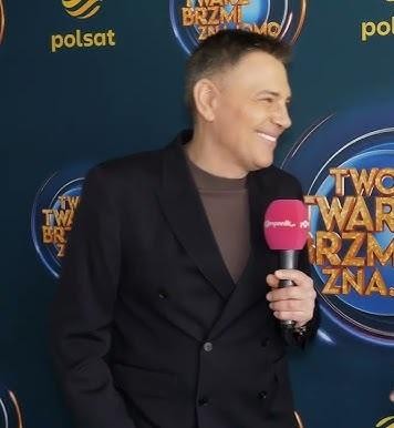 Krzysztof Ibisz prowadzący program telewizyjny w studio, uśmiechnięty i charyzmatyczny.